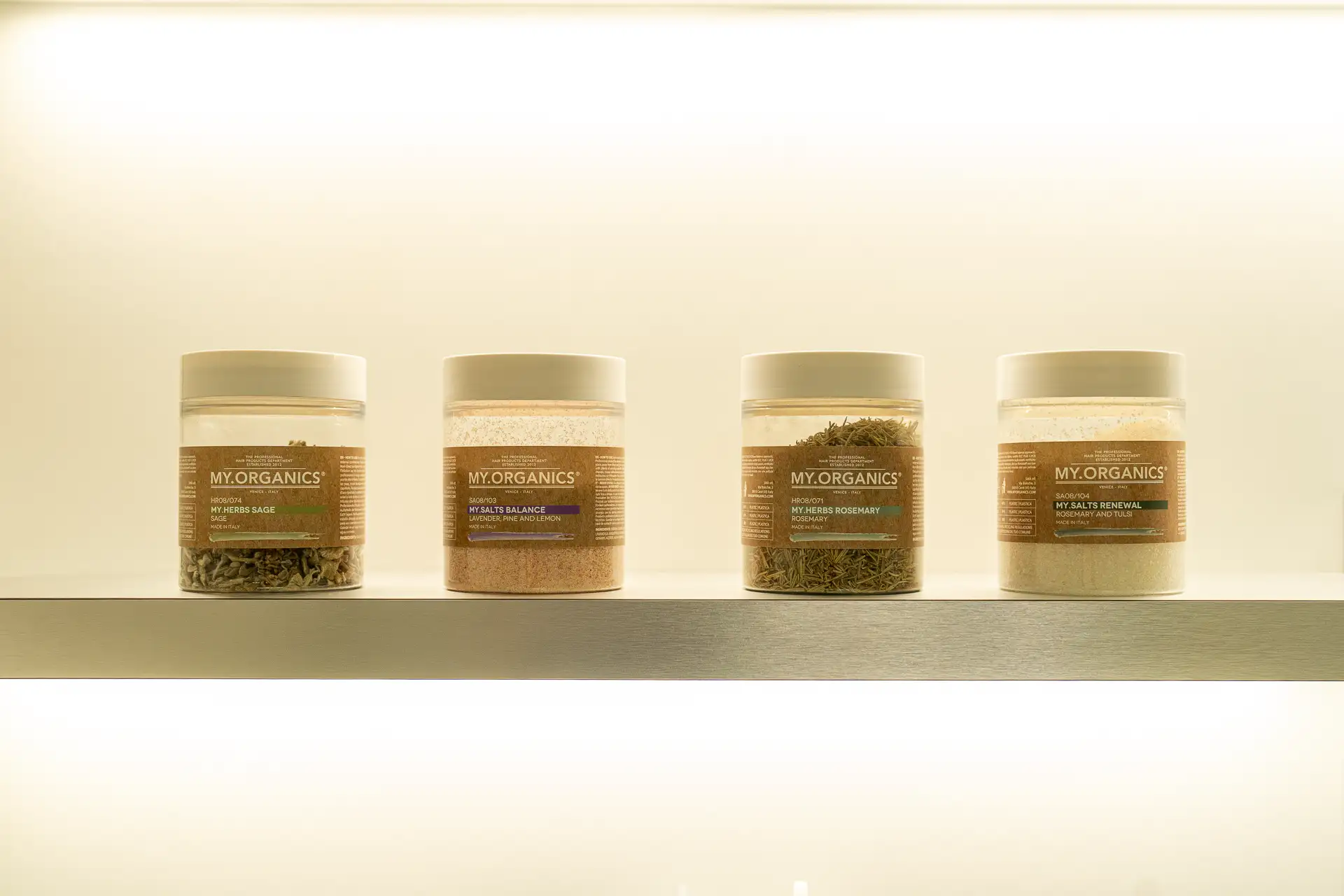Quattro barattoli My.Organics con erbe e sali su una mensola illuminata della cabina Head Spa